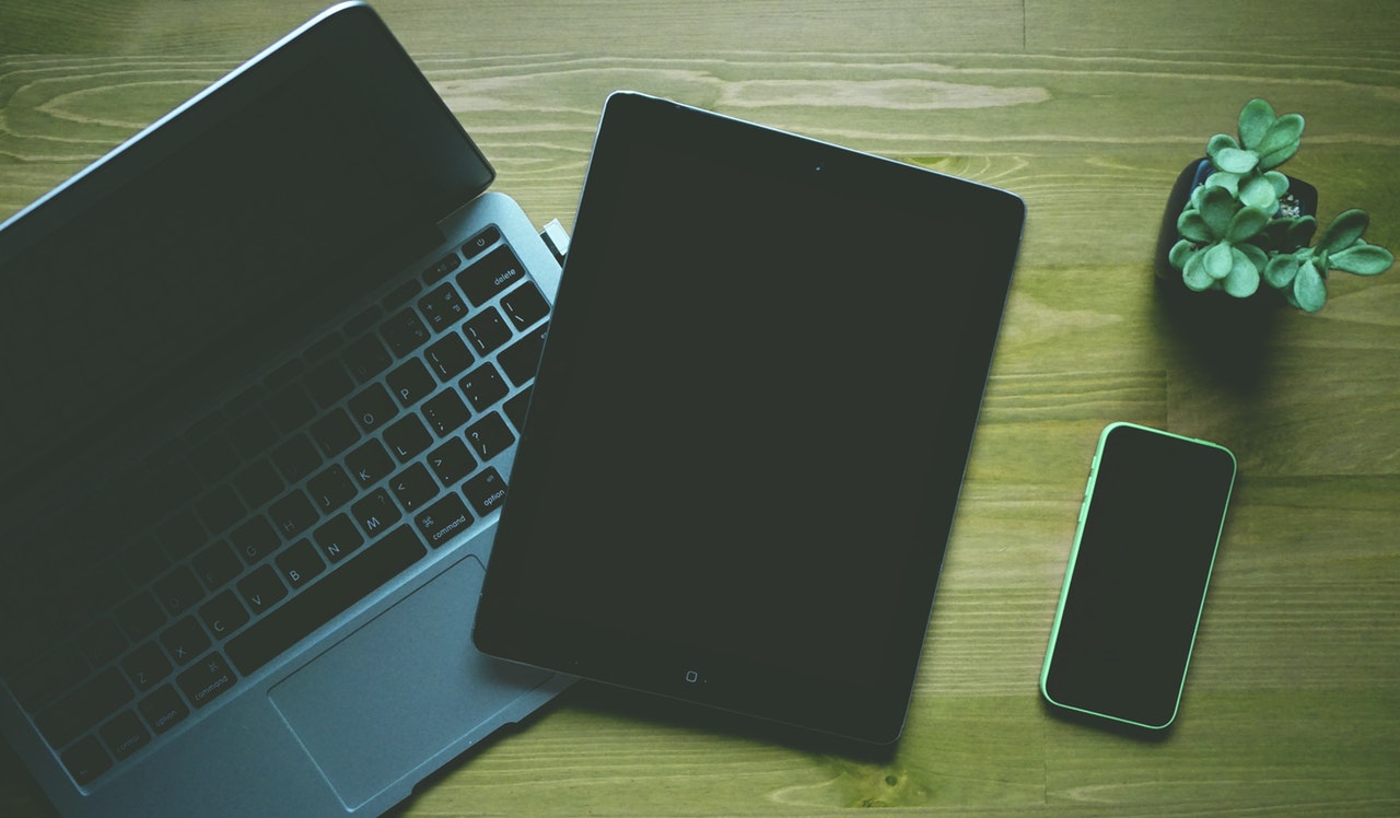 photo of a laptop, iPad, and iPhone on a wooden desk next to a plant